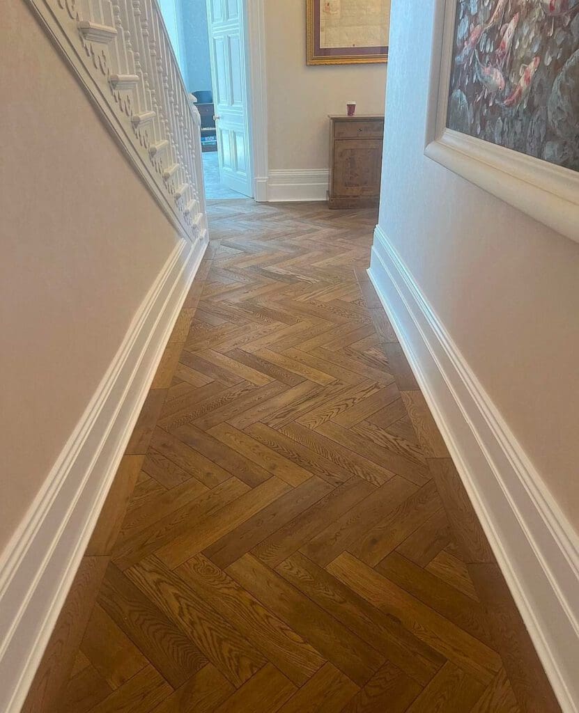 A hallway showcases a herringbone-patterned wooden floor, a testament to expert carpentry services. Light-colored walls with white baseboards are adorned with paintings, and at the hall's end, a wooden cabinet and red cup complete this inviting space.