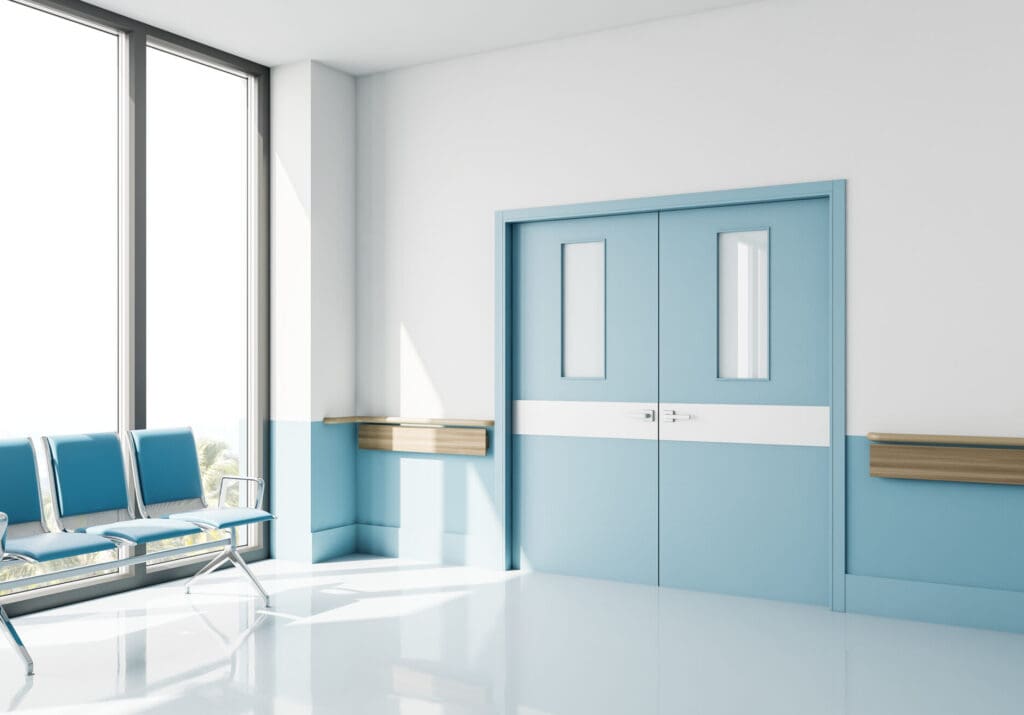 A bright hospital corridor with blue and white walls boasts expertly crafted fire door installations in the center. A row of blue chairs lines the left, while large windows let natural light illuminate the glossy floor, reflecting a commitment to both safety and aesthetics.
