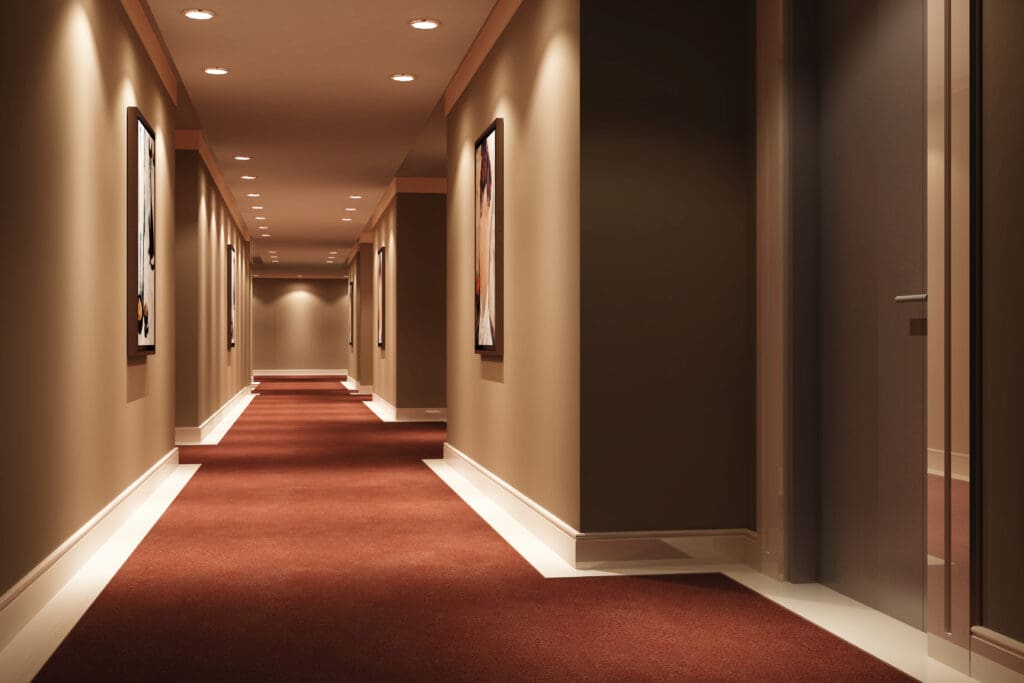 A long, well-lit hallway with brown walls and a red carpet features expertly installed fire doors on the right. The ceiling has small recessed lights, and picture frames are mounted along the walls, showcasing the craftsmanship of our carpentry services.