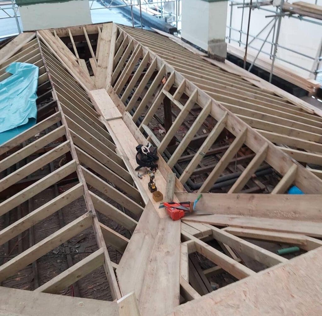A wooden roof under construction displays exposed beams and planks, hinting at ongoing home renovations. Tools like a drill and measuring tape rest on the central beam, while scaffolding encircles the structure. A blue tarp partially covers one section, adding a splash of color to the site.