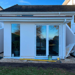 A small white extension with large glass doors and a flat roof, crafted by a building company. The doors reflect the blue sky and trees. Angular side walls, a measuring tape on the ground, and a metal tin sit by the right wall. Grass and gravel frame this sleek structure in the foreground.