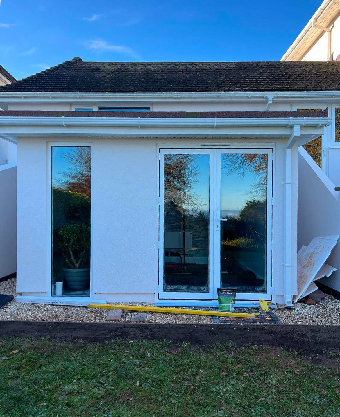 A small white extension with large glass doors and a flat roof, crafted by a building company. The doors reflect the blue sky and trees. Angular side walls, a measuring tape on the ground, and a metal tin sit by the right wall. Grass and gravel frame this sleek structure in the foreground.