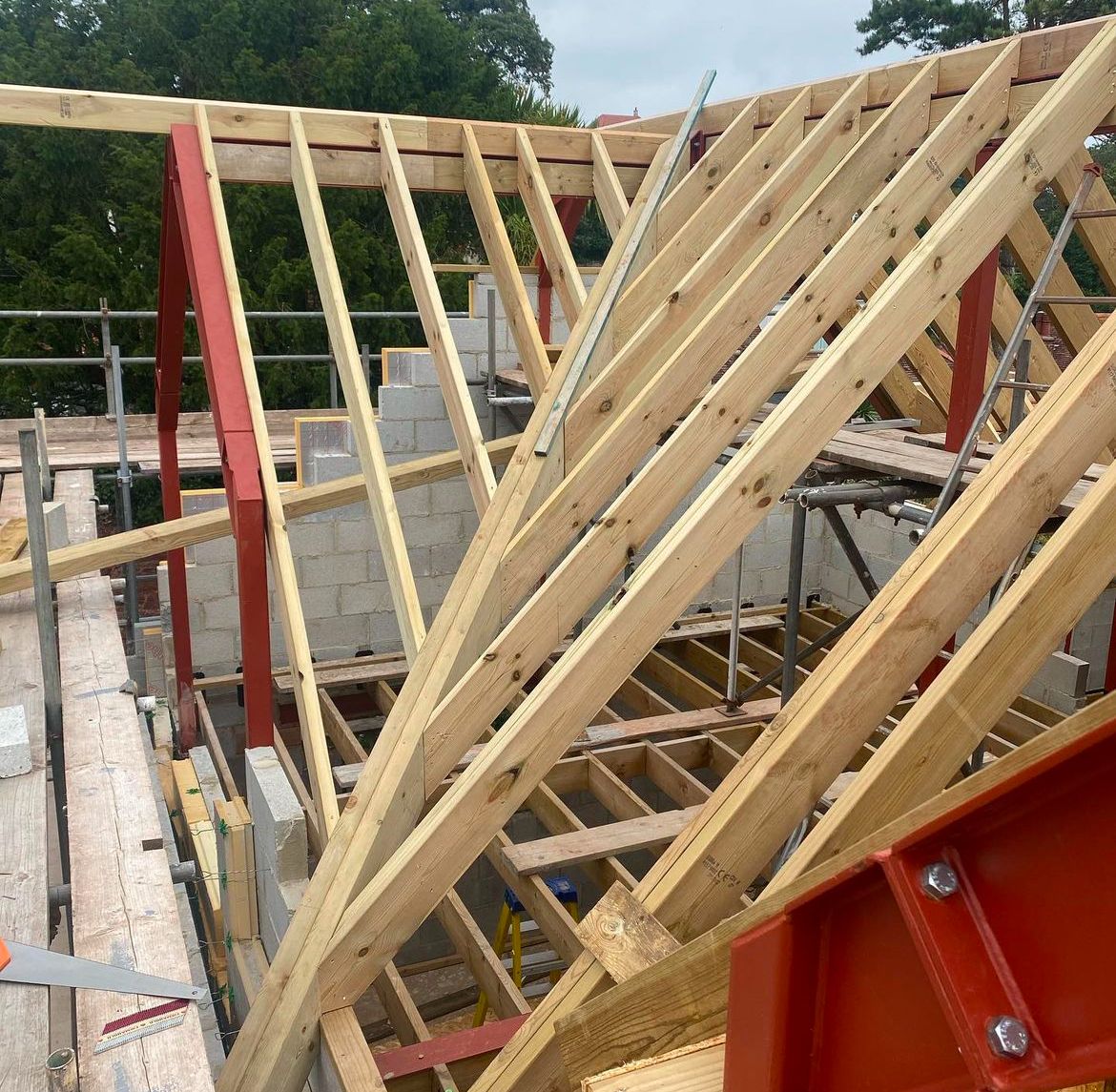 Wooden roof trusses under construction at a building site, expertly crafted by a skilled building company. Surrounded by scaffolding, the framework stands against a backdrop of trees, with cinder block walls partially visible.