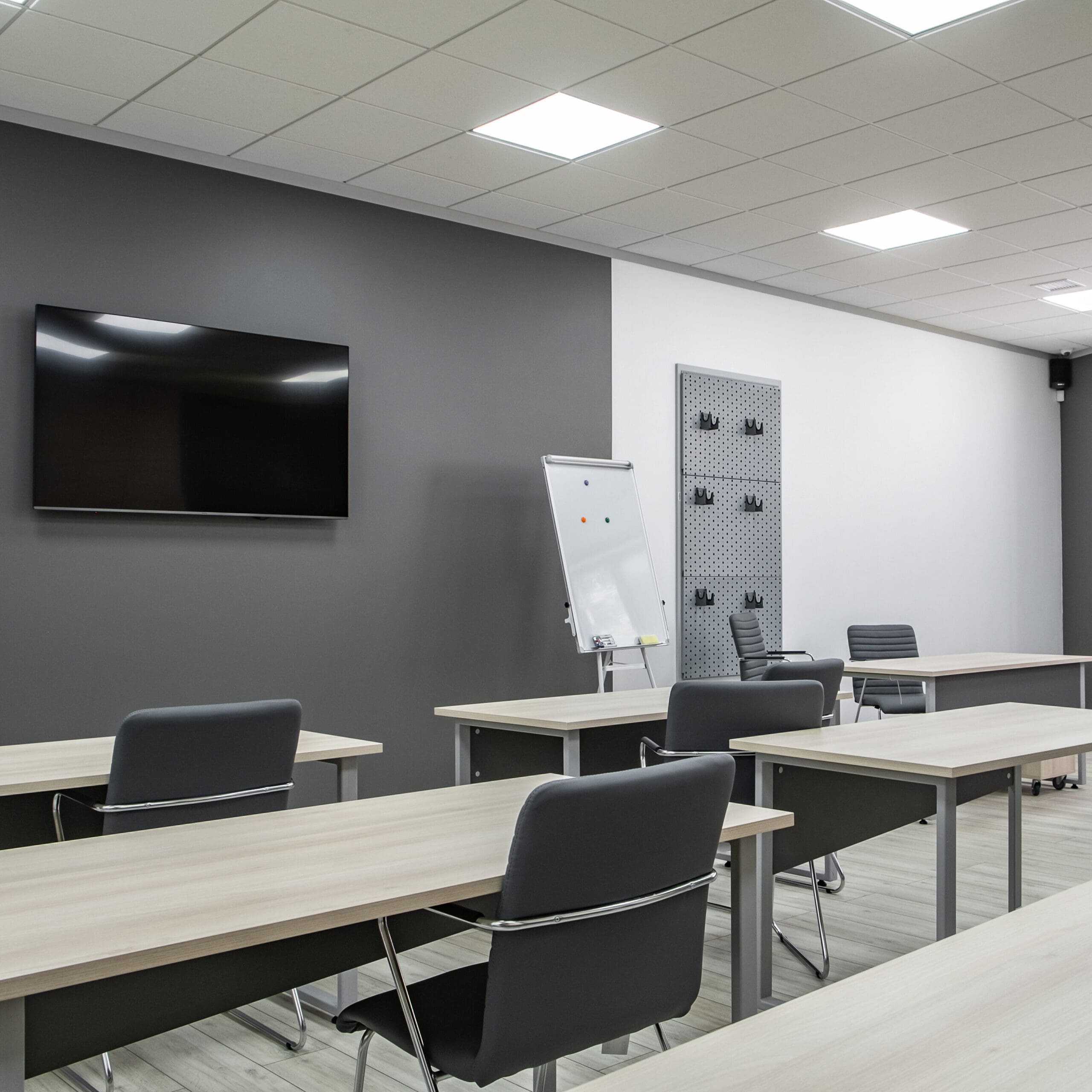 A modern, empty classroom with several wooden desks and gray chairs. A whiteboard stands near a flat-screen TV on the wall. Natural light comes through large windows with blinds partially open. The floor is light-colored wood.