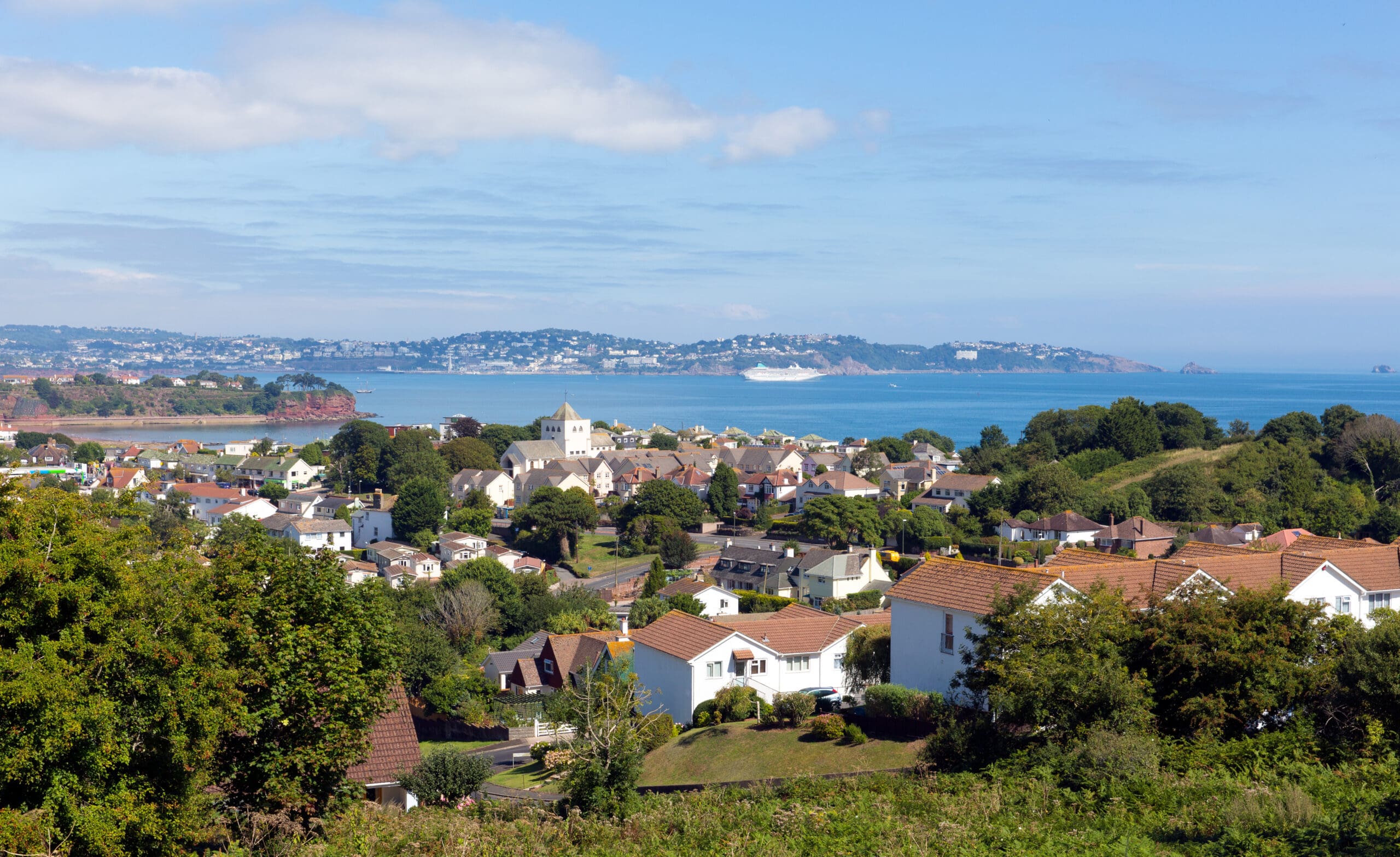 A coastal town with white houses surrounded by greenery overlooks a bay where a large ship is docked. Hills speckled with homes hint at the craftsmanship of a local building company, all beneath a blue sky decorated with scattered clouds.