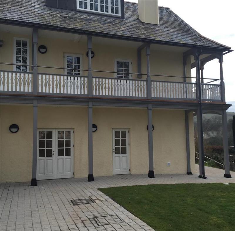 A two-story building with beige walls and a gray roof features a covered balcony on the upper floor. There are white double doors on both levels. The foreground has a tiled patio and a small patch of grass. Trees are visible in the background.